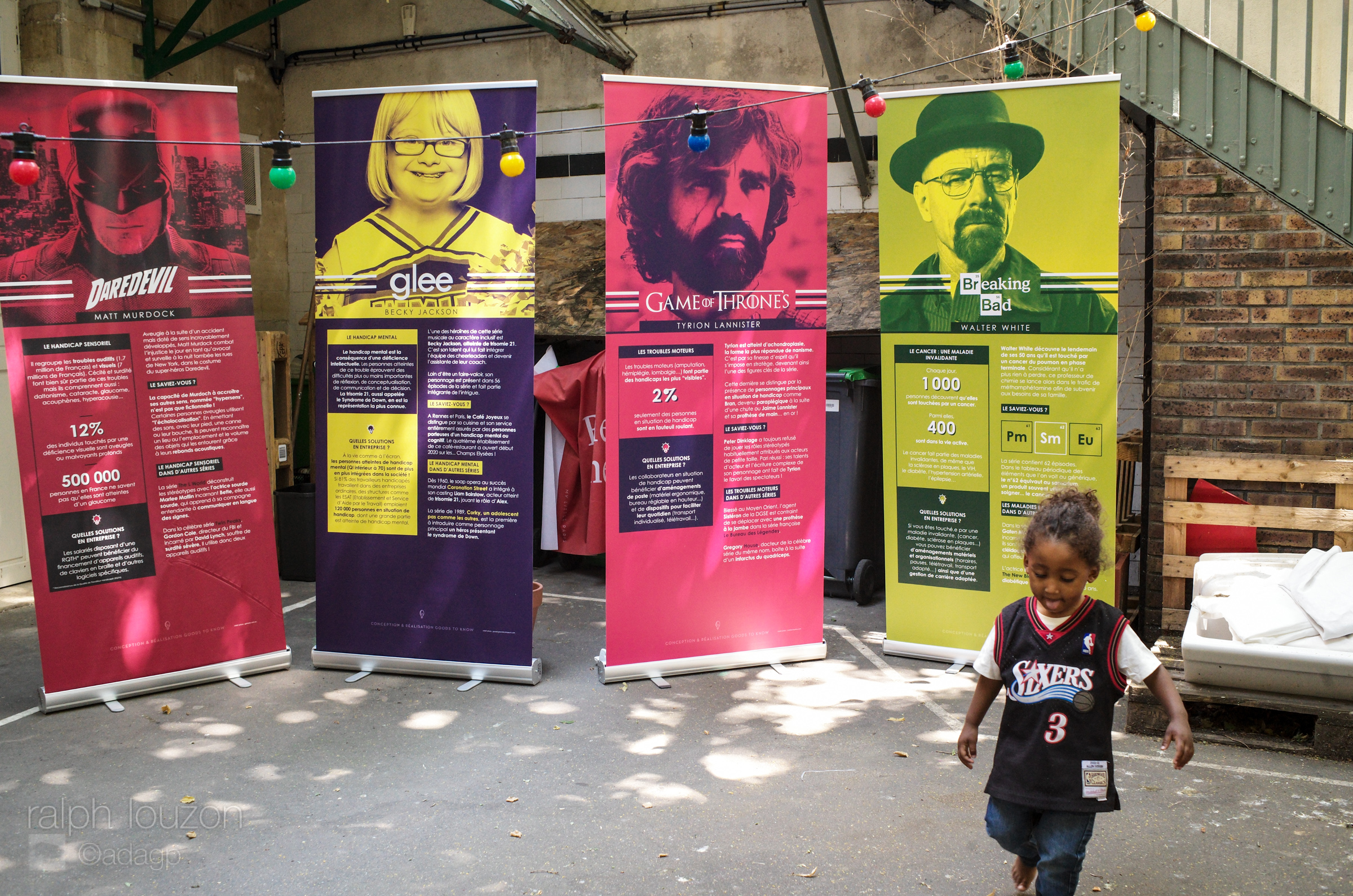 Une enfant cours au premier plan. A l'arrière plan, 4 kakémonos de l'exposition Séries et Handicap