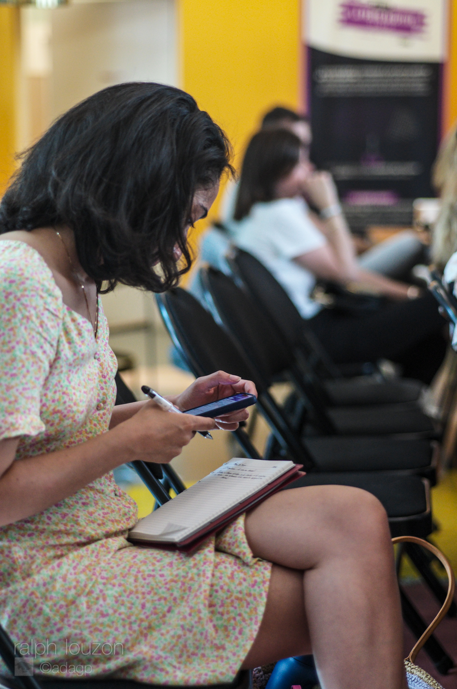 Une femme assise regarde son téléphone portable, un stylo dans la main et un carnet sur ses genoux