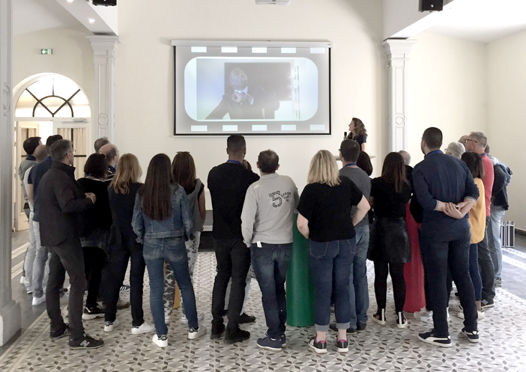 Animation Ciné quiz. Au premier plan se tient un groupe de collaborateurs, qui regardent un écran géant avec un extrait de film diffusé, en arrière plan.